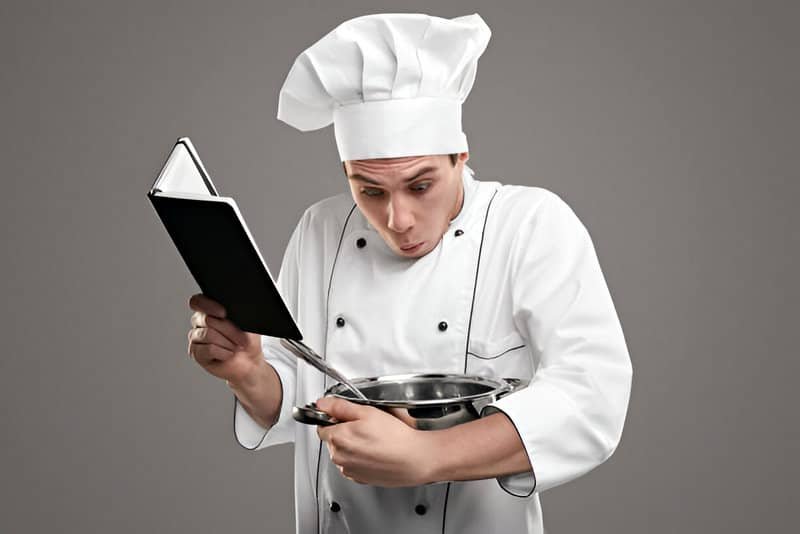 Cocinero con un libro de preguntas frecuentes en una mano y un bol en la otra, mirando hacia abajo.